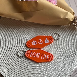 May include: Two orange keyrings with white text and nautical symbols. The keyring on the left has a steering wheel, anchor, and sailboat. The keyring on the right says "BOAT LIFE".