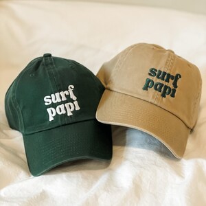 May include: Two baseball caps with the text "surf papi" embroidered on them. One cap is green and the other is tan.