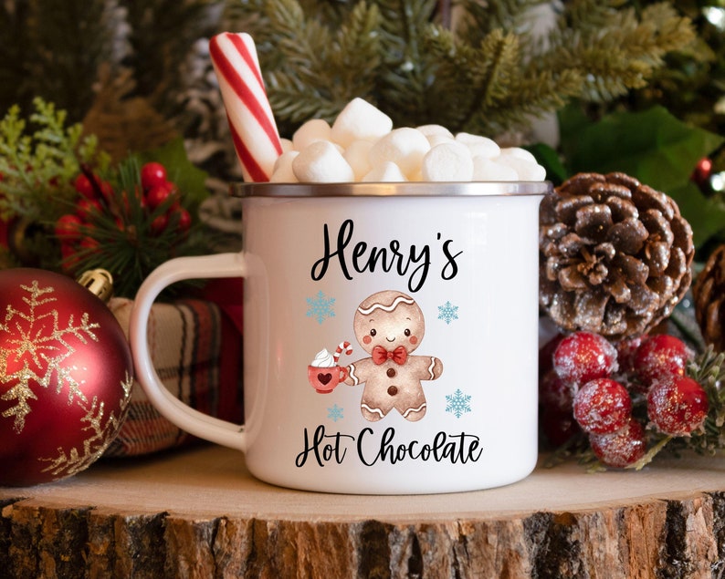 Puede incluir: Taza de esmalte blanco con un dise&ntilde;o de hombre de jengibre y el texto "Henry's Hot Chocolate". La taza est&aacute; llena de malvaviscos y un bast&oacute;n de caramelo.