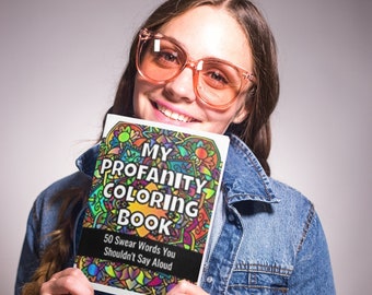 May include: A woman wearing pink glasses and a denim jacket holds a coloring book titled "My Profanity Coloring Book" with the text "50 Swear Words You Shouldn't Say Aloud".