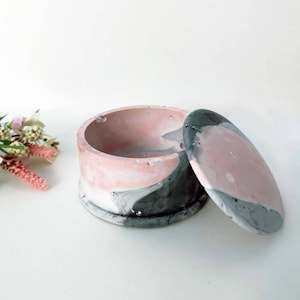 May include: A round, pink and grey concrete trinket box with a lid. The box has a marbled design.