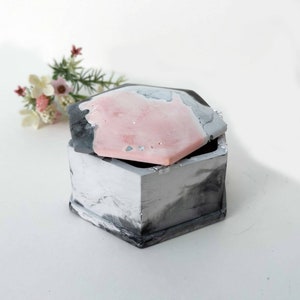 May include: A hexagonal concrete trinket box with a lid. The box is grey with black marbling and the lid is pink with grey marbling.