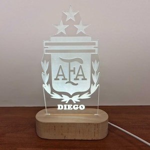 Puede incluir: Una luz nocturna de acrílico blanco con una base de madera. La luz presenta una silueta blanca del escudo de la Asociación del Fútbol Argentino (AFA) con tres estrellas encima. El nombre "DIEGO" está impreso debajo del escudo.