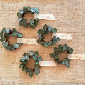 May include: Five small eucalyptus wreaths with white ribbon tied around each one. The ribbon has the text "M+S" printed on it.