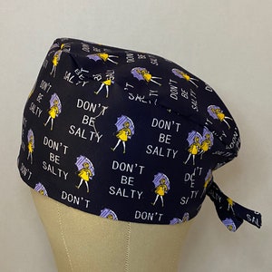May include: A black scrub hat with a repeating pattern of a woman holding an umbrella and the text "Don't Be Salty".