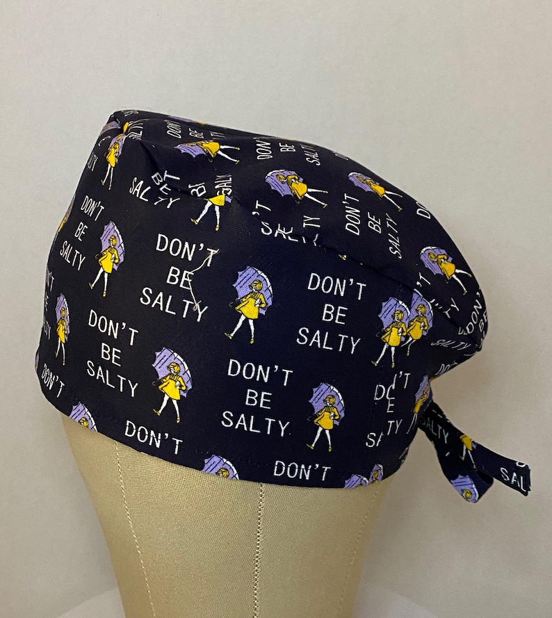 May include: A black scrub hat with a repeating pattern of a woman holding an umbrella and the text "Don't Be Salty".