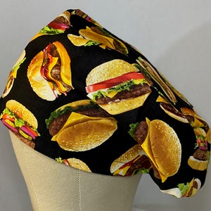 May include: A black chef's hat with a repeating pattern of colorful cartoon hamburgers. The hat is made of fabric and has a rounded shape.