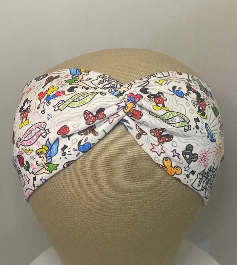 May include: White headband featuring a colourful print of various Disney characters, including Mickey Mouse, Minnie Mouse, Tinkerbell, and other iconic symbols. The headband is twisted and has a white background.