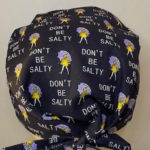 May include: Black scrub hat with a repeating pattern of a woman holding an umbrella and the text "Don't Be Salty". The hat has yellow ties.