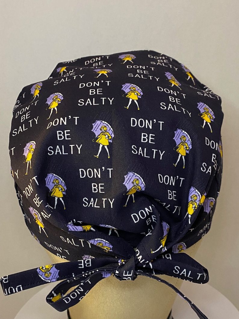 May include: Black scrub hat with a repeating pattern of a woman holding an umbrella and the text "Don't Be Salty". The hat has yellow ties.