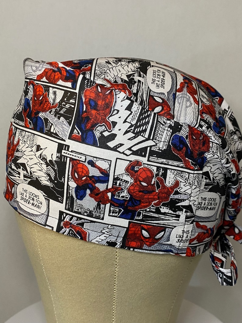 Pu&ograve; includere: Un berretto chirurgico bianco, rosso e nero con un design a fumetti con Spiderman. Il berretto ha lacci sul retro e il testo "This looks like a job for Spiderman!" &egrave; stampato sul berretto.