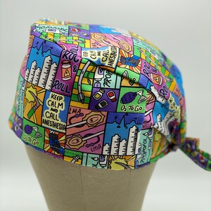 May include: A colourful surgical scrub hat with a cartoon-style print featuring medical equipment, instruments, and phrases like "Keep Calm and Call Anesthesia", "ROC & Roll", "LMA", and "O2 to Go".