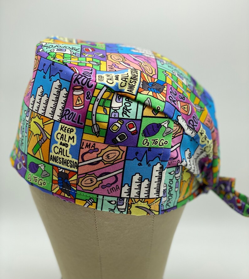 May include: A colourful surgical scrub hat with a cartoon-style print featuring medical equipment, instruments, and phrases like "Keep Calm and Call Anesthesia", "ROC & Roll", "LMA", and "O2 to Go".