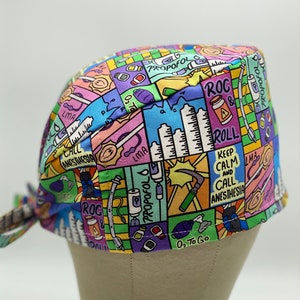 May include: A colourful surgical scrub hat with a cartoon design featuring medical equipment, medications, and phrases like "ROC & ROLL" and "Keep Calm and Call Anesthesia".
