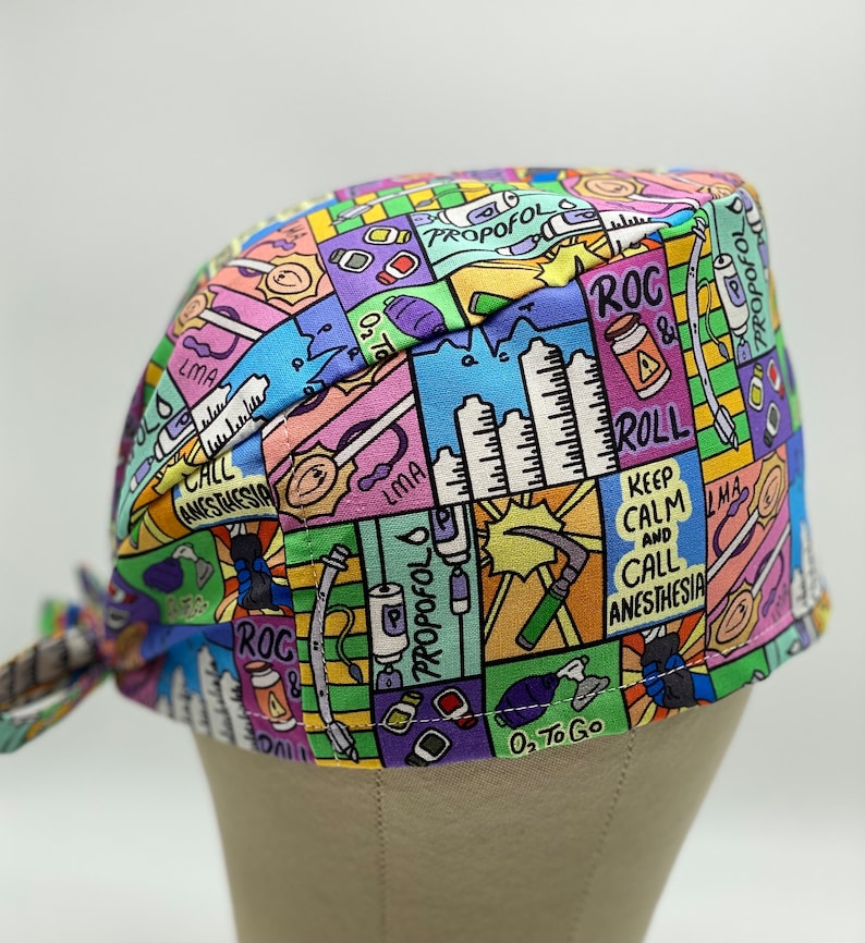 May include: A colourful surgical scrub hat with a cartoon design featuring medical equipment, medications, and phrases like "ROC & ROLL" and "Keep Calm and Call Anesthesia".