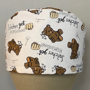 May include: White scrub hat with a brown teddy bear and the text "Snitches get stitches" repeated throughout the fabric.