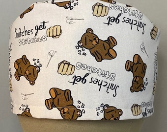 Könnte beinhalten: Eine weiße OP-Mütze mit einem braunen Teddybär und dem Text "Snitches get stitches", der sich über den gesamten Stoff wiederholt.