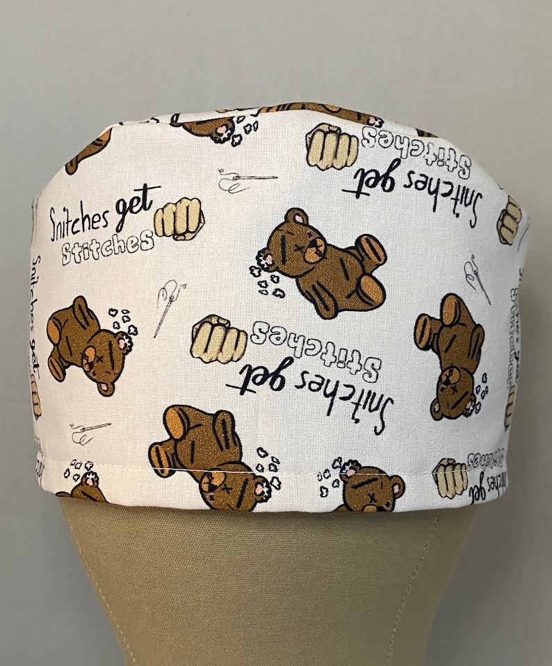 May include: White scrub hat with a brown teddy bear and the text "Snitches get stitches" repeated throughout the fabric.