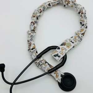 May include: A black stethoscope with a white fabric stethoscope holder featuring a pattern of dogs. The holder has a button closure.