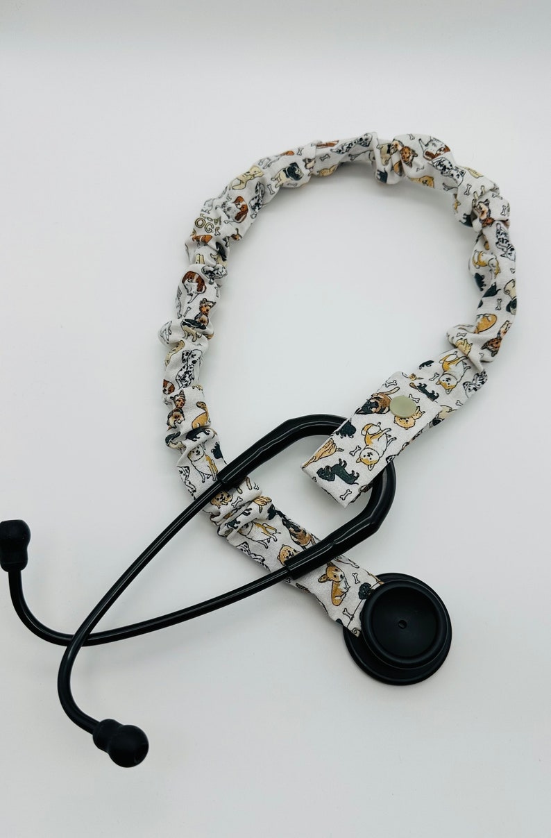 May include: A black stethoscope with a white fabric stethoscope holder featuring a pattern of dogs. The holder has a button closure.