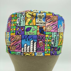 May include: A colourful surgical scrub hat with a cartoon-style design featuring medical equipment, text, and illustrations. The text includes "Keep Calm and Call Anesthesia", "ROC & Roll", "LMA", "O₂ To Go", and "Propofol".