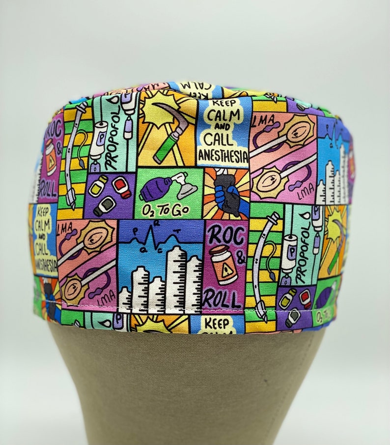 May include: A colourful surgical scrub hat with a cartoon-style design featuring medical equipment, text, and illustrations. The text includes "Keep Calm and Call Anesthesia", "ROC & Roll", "LMA", "O₂ To Go", and "Propofol".