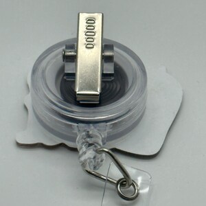 Peut inclure: Un porte-badge r&eacute;tractable en plastique transparent avec un clip argent&eacute; et un fond blanc. Le clip porte le num&eacute;ro 00000.