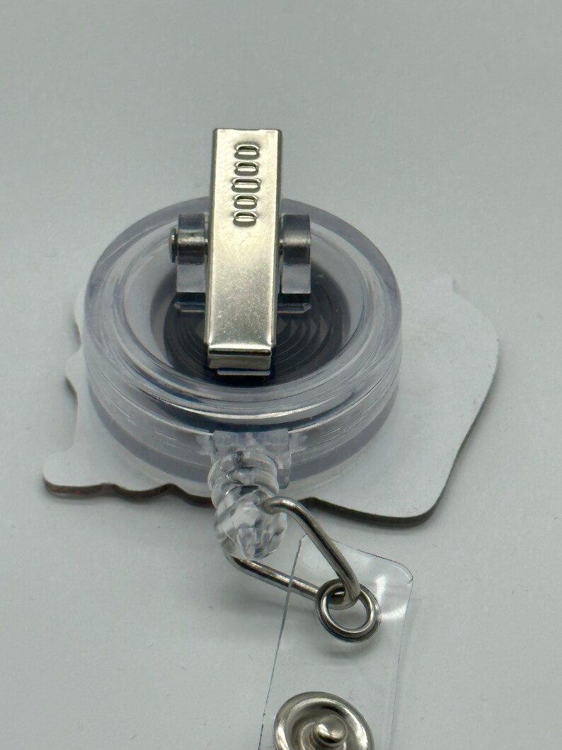 Peut inclure: Un porte-badge r&eacute;tractable en plastique transparent avec un clip argent&eacute; et un fond blanc. Le clip porte le num&eacute;ro 00000.
