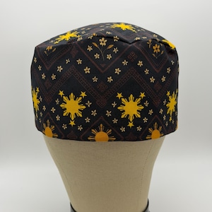 May include: A black surgical scrub cap featuring a repeating pattern of yellow suns, stars, and floral elements. The cap is displayed on a mannequin head. The design incorporates a geometric border in a reddish-brown hue.