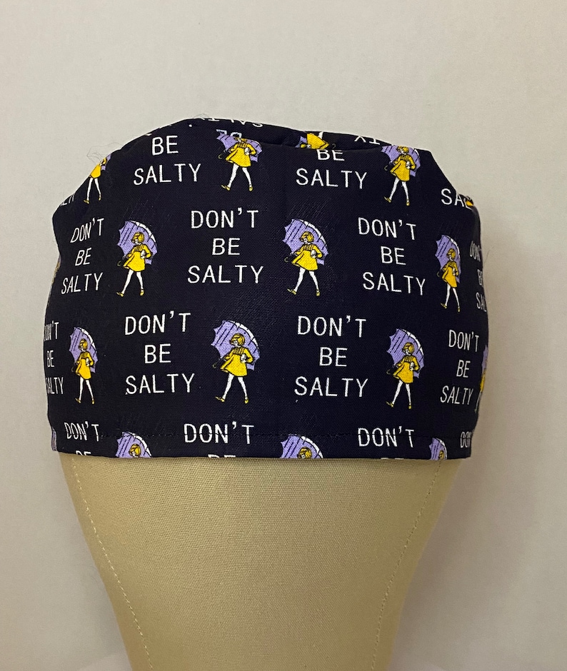 May include: A black scrub hat with a repeating pattern of yellow stick figures holding purple umbrellas and the text "Don't be salty" and "Be salty".