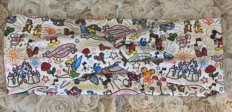 May include: White fabric headband featuring a colourful print of Disney characters, including Mickey Mouse, Minnie Mouse, Tinkerbell, and Chip and Dale. The print also includes the words "Walt Disney World" and "Disneyland".