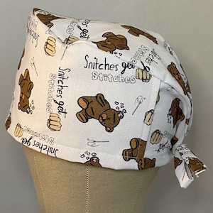 May include: White scrub hat with a brown teddy bear print and the text "Snitchers get stitches" repeated throughout.