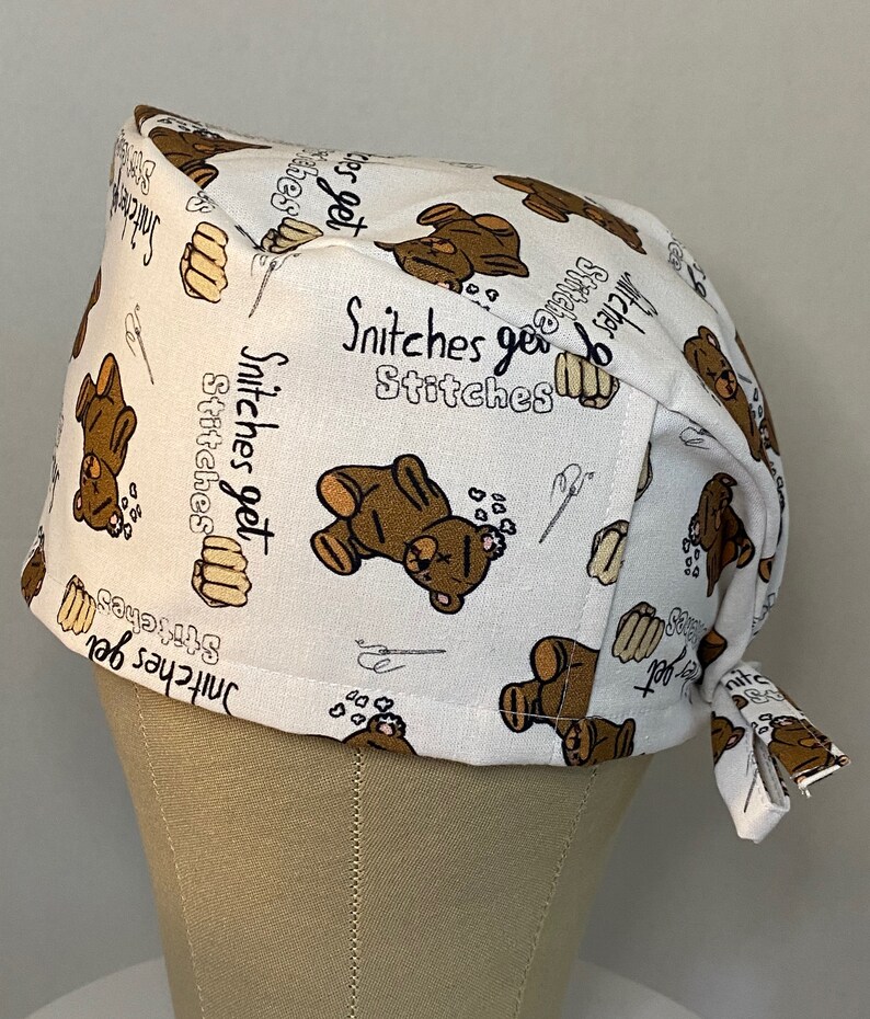 May include: White scrub hat with a brown teddy bear print and the text "Snitchers get stitches" repeated throughout.