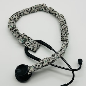 May include: A black stethoscope with a fabric cover featuring a repeating pattern of US one hundred dollar bills. The stethoscope has a black chest piece, earpieces, and tubing. The fabric cover adds a unique and stylish touch to the medical instrument.