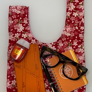 Puede incluir: Una bolsa de tela roja con estampado floral con flores rosas. La bolsa est&aacute; llena de una peque&ntilde;a libreta naranja, unas gafas marrones, un bol&iacute;grafo negro y una peque&ntilde;a botella de desinfectante de manos.