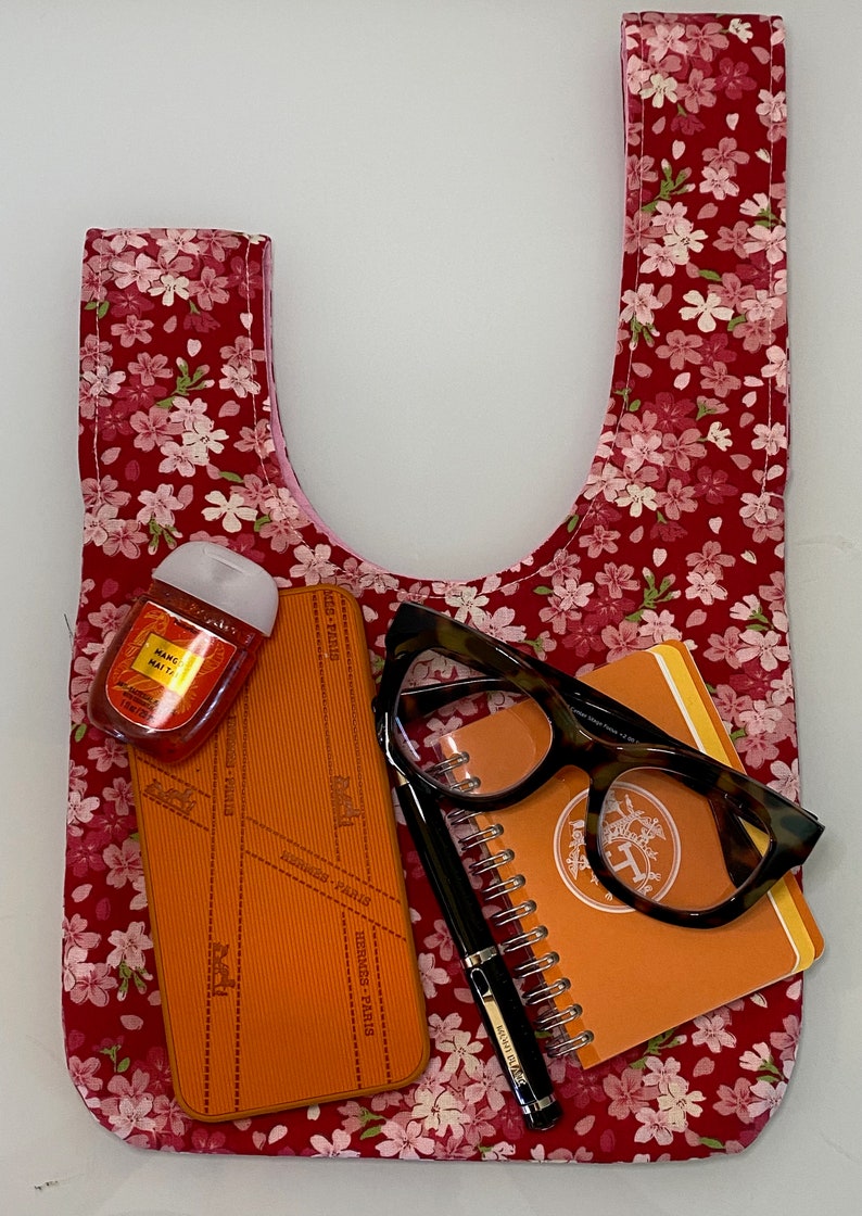 Puede incluir: Una bolsa de tela roja con estampado floral con flores rosas. La bolsa est&aacute; llena de una peque&ntilde;a libreta naranja, unas gafas marrones, un bol&iacute;grafo negro y una peque&ntilde;a botella de desinfectante de manos.