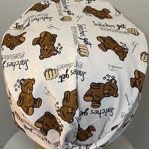 May include: White fabric scrub hat with a brown teddy bear print and the text "Snitches get stitches" repeated throughout.