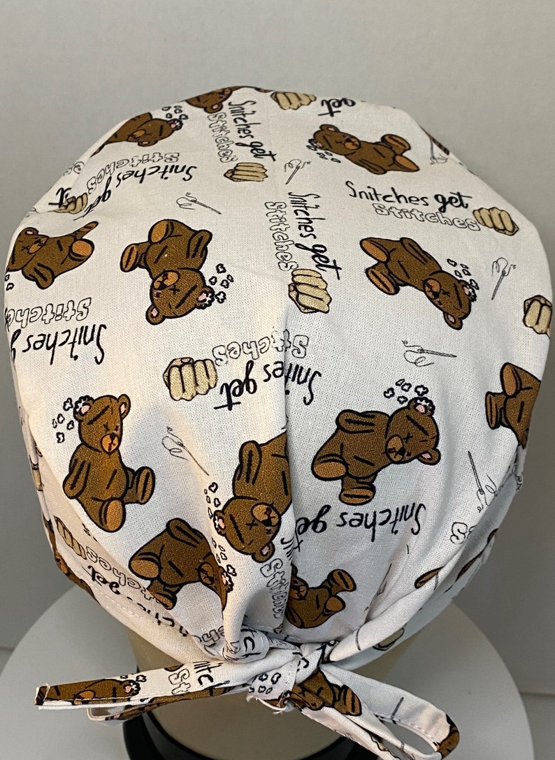May include: White fabric scrub hat with a brown teddy bear print and the text "Snitches get stitches" repeated throughout.
