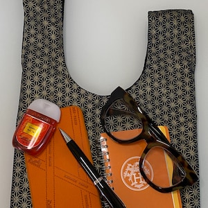 May include: A black and gold patterned tote bag with a small orange notebook, a pen, a pair of glasses, and a small bottle of hand sanitizer inside. The notebook has the Hermes logo on the front.