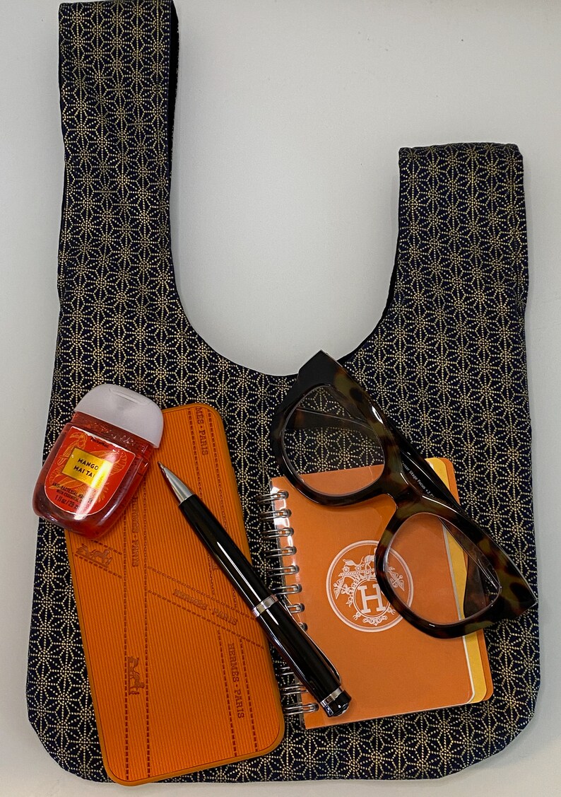 May include: A black and gold patterned tote bag with a small orange notebook, a pen, a pair of glasses, and a small bottle of hand sanitizer inside. The notebook has the Hermes logo on the front.