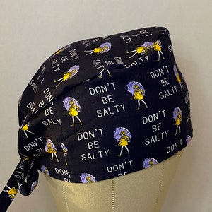 May include: Black scrub hat with a repeating pattern of a girl holding an umbrella and the text "Don't Be Salty".