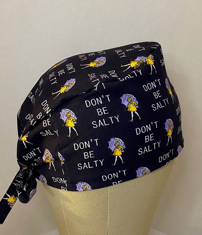 May include: Black scrub hat with a repeating pattern of a girl holding an umbrella and the text "Don't Be Salty".