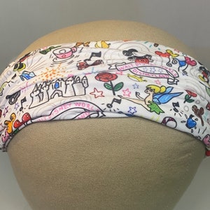 May include: White headband with a colourful print of Disney characters and symbols, including Mickey Mouse, Tinkerbell, and Cinderella's castle. The text "Walt Disney World" is visible on the headband.