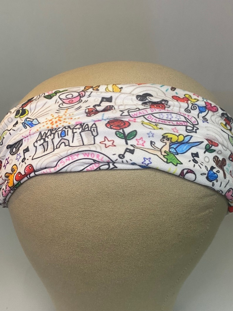 May include: White headband with a colourful print of Disney characters and symbols, including Mickey Mouse, Tinkerbell, and Cinderella's castle. The text "Walt Disney World" is visible on the headband.