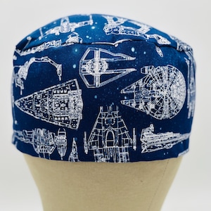 May include: A navy blue surgical scrub cap featuring a white schematic design of various spaceships. The cap is made of fabric and is displayed on a mannequin head against a white background. The design includes the Millennium Falcon and other iconic spacecraft.