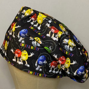 May include: Black scrub hat with a colorful pattern of M&M's characters and the text "Sweet & Nutty".