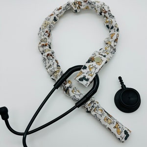May include: A black stethoscope with a white fabric cover featuring a pattern of dogs and bones. The fabric cover is attached to the stethoscope with a black clip. The stethoscope has a black diaphragm and a black earpiece.