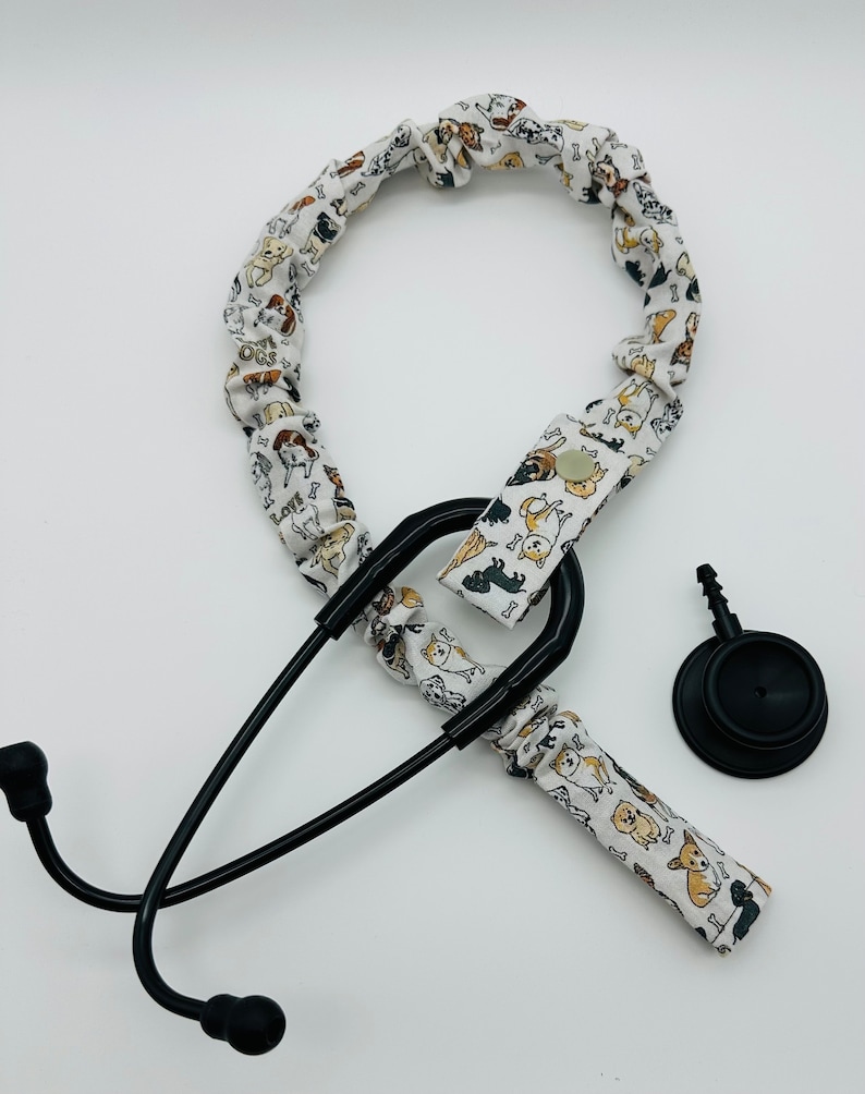 May include: A black stethoscope with a white fabric cover featuring a pattern of dogs and bones. The fabric cover is attached to the stethoscope with a black clip. The stethoscope has a black diaphragm and a black earpiece.