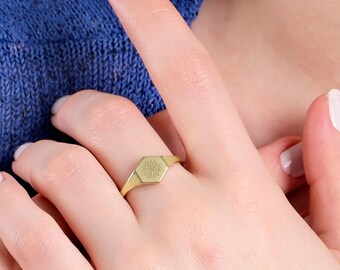 May include: A gold-toned signet ring featuring a flat, hexagonal face with a subtle, etched botanical design. The ring has a smooth, polished band and is worn on a finger, showcasing the metallic finish and the geometric shape of the centerpiece.