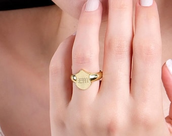 Puede incluir: Un anillo de sello de oro con una cara en forma de escudo que presenta las letras grabadas EM. El anillo tiene un acabado metálico pulido y una banda lisa y redondeada que se ajusta al dedo, mostrando un diseño clásico y sencillo.